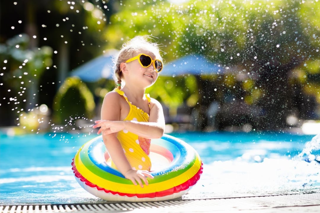 Child wearing heart-shaped sunglasses smiling in a swimming pool with a rainbow inflatable ring